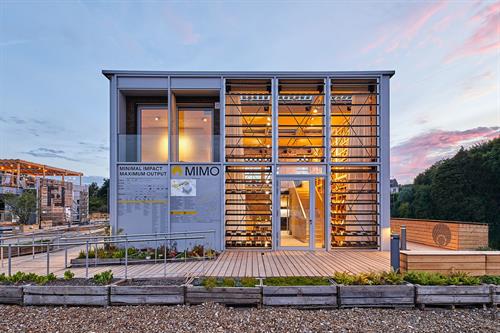Modernes, zweigeschossiges Demonstrationsgebäude „MIMO“ mit großer Glasfassade und Holzterrasse im Außenbereich bei Abendlicht.